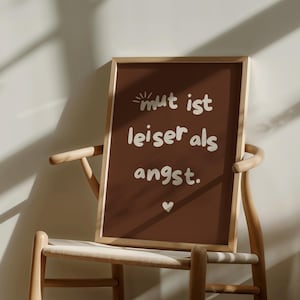 Op de afbeelding: Een ingelijste print met de Duitse tekst "mut ist leiser als angst" in witte letters op een bruine achtergrond. Onderaan staat een klein wit hartje. De lijst is lichtbruin en de print staat op een houten stoel.