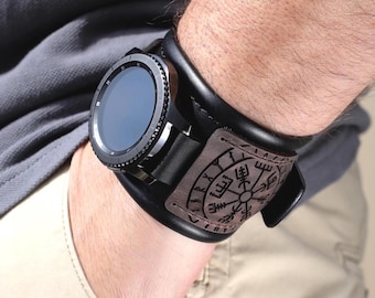 May include: Black smartwatch with a round face and a black leather cuff. The cuff features a brown leather patch with a compass rose design and Norse runes. The watch is worn on a wrist.