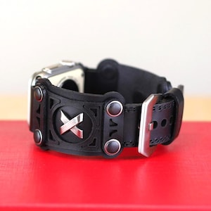 May include: Black leather watch strap with a silver buckle and rivets. The strap has a silver "X" design in the centre and is attached to a silver watch face. The watch strap is displayed on a red surface.