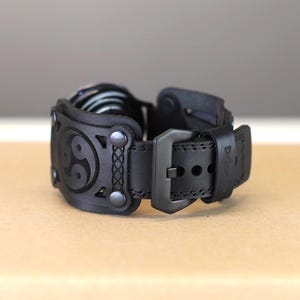 May include: Black leather watch band with a black buckle and a decorative design featuring a yin yang symbol.