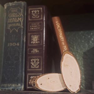 May include: A wooden shelf holds three vintage books: "The Girl's Realm Annual 1904", "Works of Charles Dickens", and "A Dictionary of Tolkien". Two oval wooden coasters are placed in front, displaying the natural wood grain.