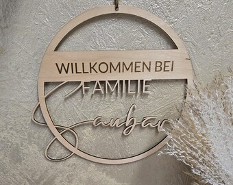 Puede incluir: Un letrero redondo de madera con las palabras "WILLKOMMEN BEI FAMILIE" y un nombre en cursiva, colgado por un lazo de cuerda. El letrero es de madera clara y está contra una pared texturizada.