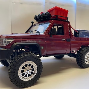 Pode incluir: Um camião de brinquedo de cor bordô com pneus off-road grandes e jantes prateadas. Possui um recipiente de armazenamento vermelho no tejadilho e um pára-choques dianteiro preto. As palavras "Land Cruiser" são visíveis na lateral.