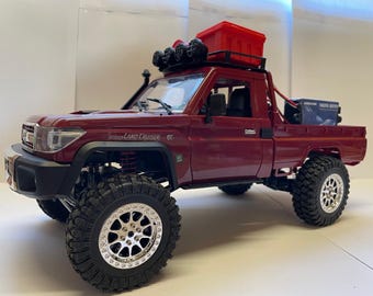 May include: A maroon-colored toy truck with large, off-road tires and silver rims. The truck has a red storage container on the roof and a black front bumper. The words "Land Cruiser" are visible on the side.
