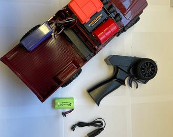 May include: A maroon-colored remote control truck with an open bed, featuring a black remote control, a green battery, and a charging cable. The truck has an orange box and a red cylinder on the bed. The remote control has a wheel and a trigger.