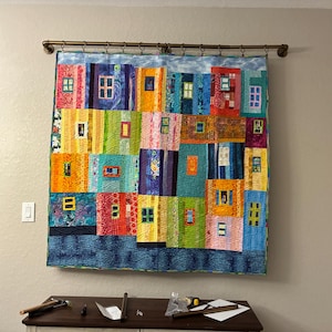 May include: A colorful, handmade quilt depicting a cityscape with rows of vibrant, rectangular windows. The quilt features a variety of colors including blue, orange, green, and red. It is hung on a wall with a decorative rod.