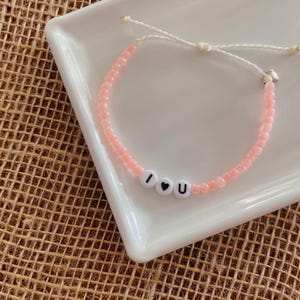 Könnte beinhalten: Ein rosa Perlenarmband mit einem weißen Kordelverschluss und weißen Buchstabenperlen, die "Ich liebe dich" buchstabieren.