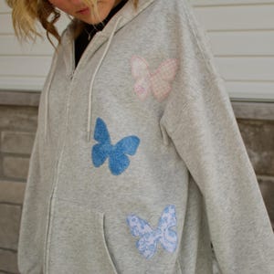 Peut inclure: Un sweat à capuche gris avec trois patchs en forme de papillon. Les papillons sont roses, bleus et blancs avec des motifs floraux.