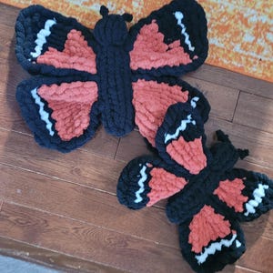 Op de afbeelding: Twee gehaakte vlinderdecoraties in zwart en oranje met witte accenten. De grotere vlinder ligt bovenop de kleinere.