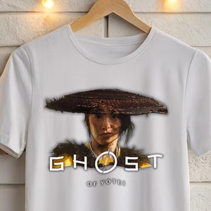 Może przedstawiać: Biała koszulka z grafiką przedstawiającą osobę w tradycyjnym kapeluszu, ze słowem "GHOST" w stylizowanej czcionce. Pod słowem "GHOST" znajduje się tekst "OF TSUSHIMA". Koszulka wisi na drewnianym wieszaku.