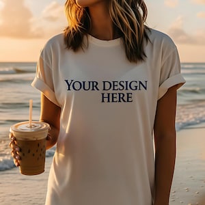 Può includere: T-shirt color crema con maniche arrotolate e la scritta "YOUR DESIGN HERE" in blu navy. La persona tiene in mano un bicchiere di plastica trasparente con caffè freddo e una cannuccia. Lo sfondo mostra una spiaggia e l'oceano.