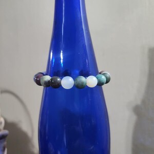 May include: A bracelet made of round, multi-colored gemstone beads. The beads are a mix of green, brown, white, and gray.