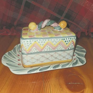 May include: A ceramic butter dish with a lid, resting on a matching tray. The dish is rectangular with a checkered yellow and white top, and a decorative band of pink, green, and yellow. The handle is twisted with yellow spheres on each end.