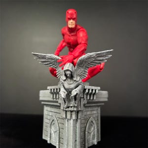 Può includere: Una action figure rossa di Daredevil è appollaiata su un diorama di tetto grigio e dettagliato. La figura è in posizione accovacciata, con una mano alzata. Il diorama presenta un angelo scolpito con le ali.