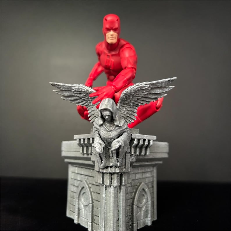 Pode incluir: Uma figura de a&ccedil;&atilde;o vermelha do Daredevil est&aacute; empoleirada em um diorama de telhado cinza e detalhado. A figura est&aacute; em uma pose agachada, com uma m&atilde;o levantada. O diorama apresenta um anjo esculpido com asas.