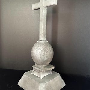 May include: A gray concrete cross with a round base and a textured sphere in the center. The cross is mounted on a square base.