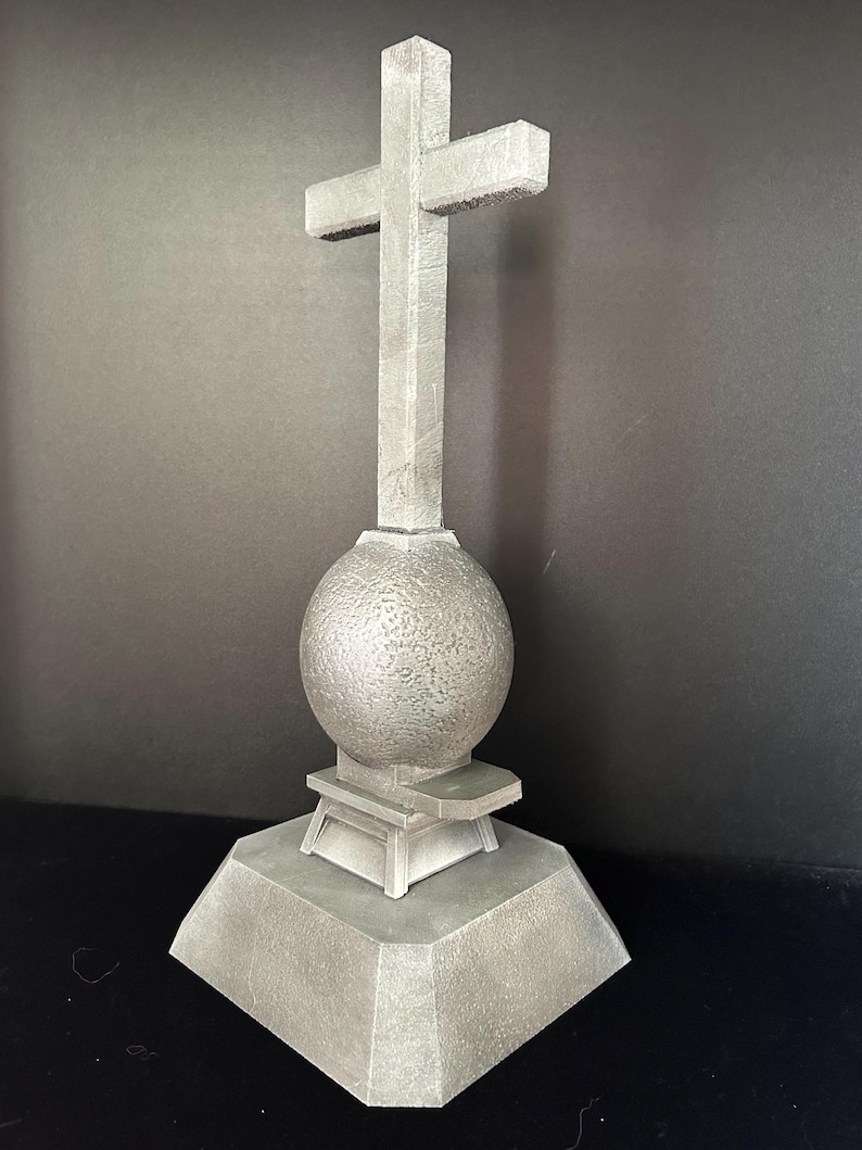 May include: A gray concrete cross with a round base and a textured sphere in the center. The cross is mounted on a square base.