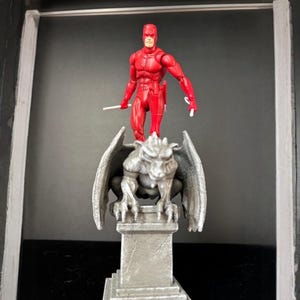 K&ouml;nnte beinhalten: Eine rote Actionfigur eines Superhelden, die zwei wei&szlig;e St&auml;be h&auml;lt, steht auf einer grauen Gargoyle-Statue. Der Superheld tr&auml;gt einen roten Anzug und eine Maske. Der Gargoyle hat detaillierte Fl&uuml;gel und sitzt auf einem quadratischen Sockel.