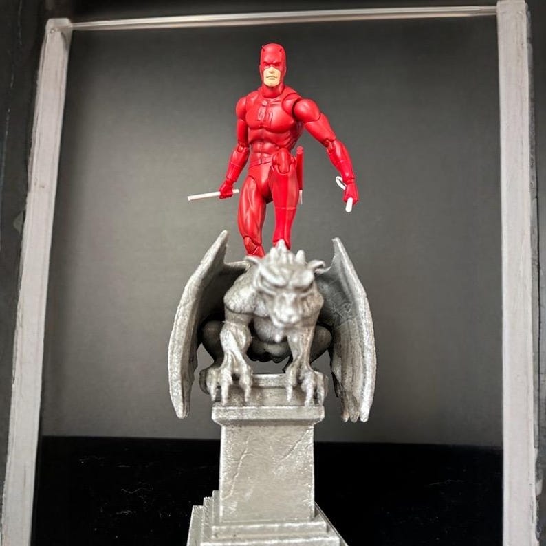 K&ouml;nnte beinhalten: Eine rote Actionfigur eines Superhelden, die zwei wei&szlig;e St&auml;be h&auml;lt, steht auf einer grauen Gargoyle-Statue. Der Superheld tr&auml;gt einen roten Anzug und eine Maske. Der Gargoyle hat detaillierte Fl&uuml;gel und sitzt auf einem quadratischen Sockel.