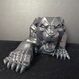 Può includere: Una scultura grigio metallizzato di una creatura felina ringhiante, forse una pantera o un leone, con dettagli precisi. La scultura è in posizione di strisciamento, con una zampa protesa in avanti. La bocca della creatura è aperta, rivelando i denti.
