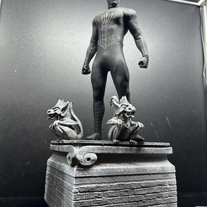 Pode incluir: Uma figura de a&ccedil;&atilde;o preta do Homem-Aranha est&aacute; em cima de uma base de pedra cinza com duas g&aacute;rgulas. A figura est&aacute; em uma pose din&acirc;mica, com um padr&atilde;o de teia detalhado no traje. A base tem uma textura semelhante a tijolos.