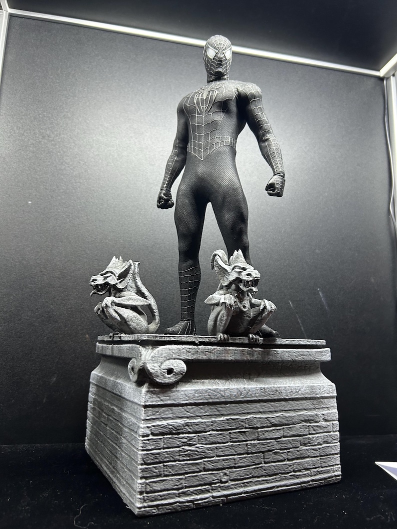 Pode incluir: Uma figura de a&ccedil;&atilde;o preta do Homem-Aranha est&aacute; em cima de uma base de pedra cinza com duas g&aacute;rgulas. A figura est&aacute; em uma pose din&acirc;mica, com um padr&atilde;o de teia detalhado no traje. A base tem uma textura semelhante a tijolos.