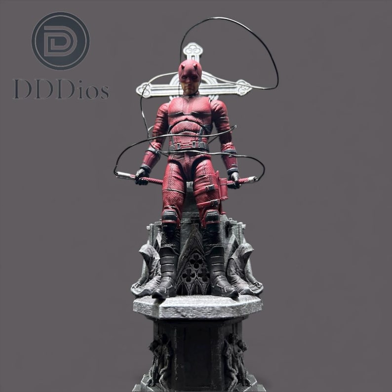 May include: A red Daredevil action figure stands atop a detailed gray pedestal. The figure is in a dynamic pose, holding two batons and a whip. The figure is wearing a textured red suit with black accents.