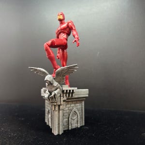 Pode incluir: Uma figura de a&ccedil;&atilde;o de super-her&oacute;i vermelha em p&eacute; sobre uma base de pedra cinza com uma est&aacute;tua de g&aacute;rgula. O super-her&oacute;i est&aacute; em pose com uma perna levantada e um bra&ccedil;o estendido.