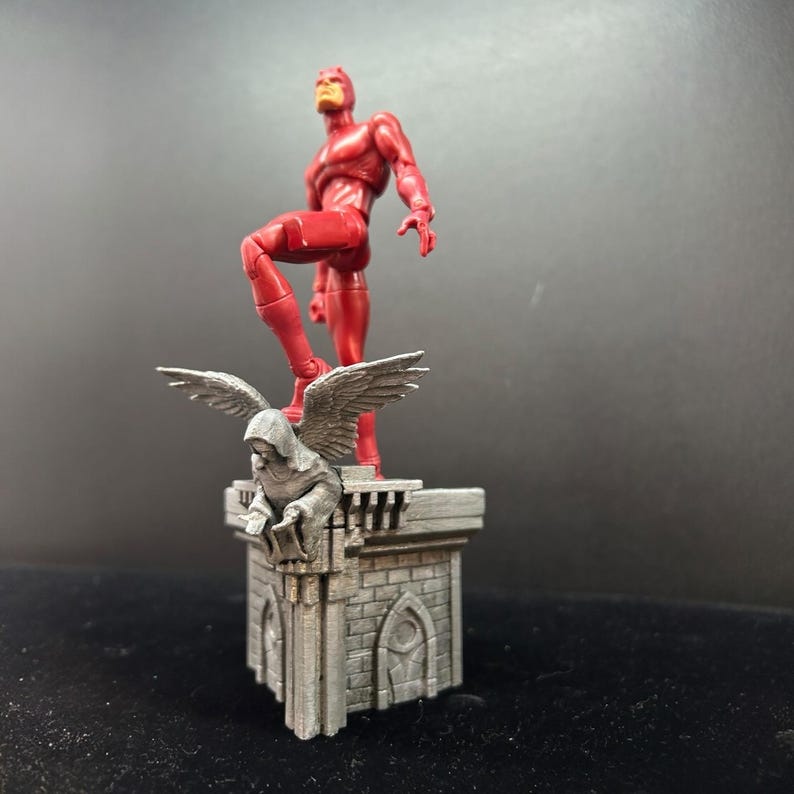 Pode incluir: Uma figura de a&ccedil;&atilde;o de super-her&oacute;i vermelha em p&eacute; sobre uma base de pedra cinza com uma est&aacute;tua de g&aacute;rgula. O super-her&oacute;i est&aacute; em pose com uma perna levantada e um bra&ccedil;o estendido.