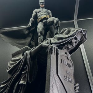 Può includere: Una statua dettagliata di Batman in abito grigio, mantello e cappuccio, in piedi su una figura della Morte. La Morte tiene una lapide con le parole "PROTECT AND SERVE" incise. La statua è ambientata su uno sfondo scuro.