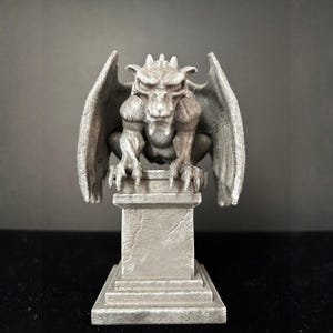 K&ouml;nnte beinhalten: Eine graue Gargoyle-Statue mit detaillierten Fl&uuml;geln, Krallen und einem grimmigen Gesichtsausdruck. Die Statue thront auf einem quadratischen Sockel und zeigt einen gotischen Architekturstil. Die Skulptur besteht aus einem stein&auml;hnlichen Material.
