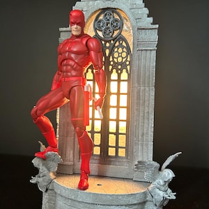 Può includere: Action figure rossa di un supereroe, in posa su una piattaforma grigia ornata con statue di angeli. Dietro la figura c'è una finestra grigia in stile gotico con luce. La figura tiene in mano un oggetto bianco. Collezionismo.