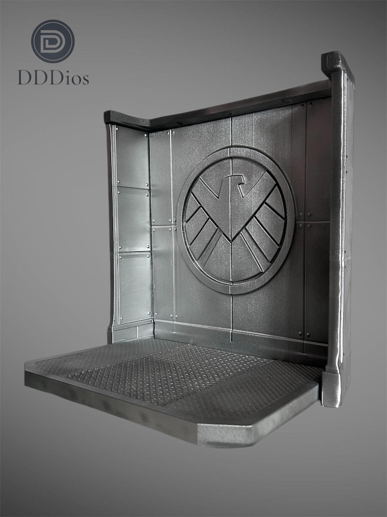 May include: A silver-colored display stand with a textured platform and a wall featuring a circular emblem with a stylized bird design. The stand has a corner design and is made of metal. The text "DDDios" is in the upper left corner.