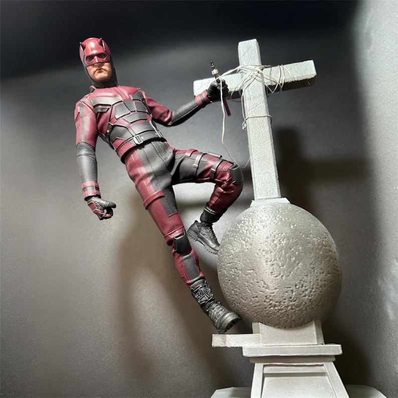 May include: A red and black action figure of Daredevil standing on a gray stone base with a cross. The figure is posed in a dynamic action pose with one leg raised and the other foot resting on the base. The figure is holding a red weapon in one hand.