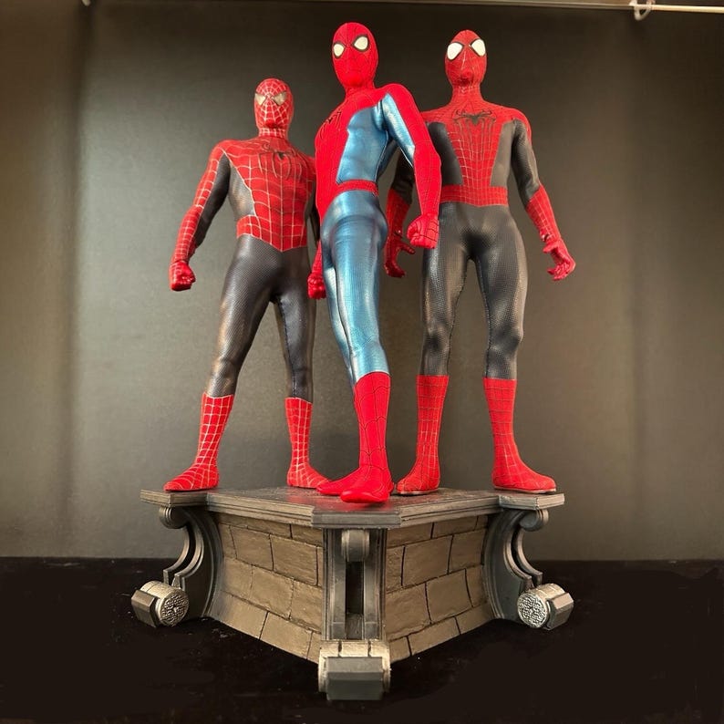 May include: Three action figures of Spider-Man in different poses standing on a stone base. The figures are all wearing red and blue suits with black webbing.