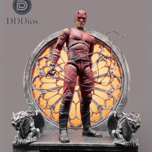 Può includere: Action figure rossa di un supereroe con le corna, in piedi davanti a un design di vetrata. La figura indossa un abito rosso e stivali neri. La base presenta statue di gargoyle su ogni lato.