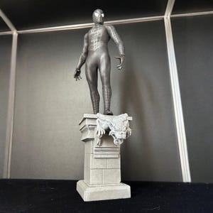 Può includere: Una statua grigia di Spider-Man è in piedi su un piedistallo di pietra con un gargoyle. La figura è in una posa classica, con un costume dettagliato. La statua è un oggetto da collezione.