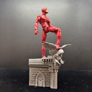 Pode incluir: Uma figura de a&ccedil;&atilde;o vermelha do Daredevil em p&eacute; em um telhado cinza com uma est&aacute;tua de g&aacute;rgula. A g&aacute;rgola tem asas e est&aacute; empoleirada na borda do telhado.