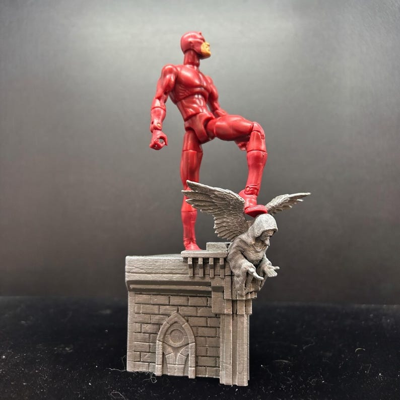 Pode incluir: Uma figura de a&ccedil;&atilde;o vermelha do Daredevil em p&eacute; em um telhado cinza com uma est&aacute;tua de g&aacute;rgula. A g&aacute;rgola tem asas e est&aacute; empoleirada na borda do telhado.