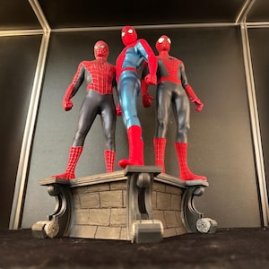May include: Three red and blue Spider-Man action figures stand on a gray stone base. The figures are posed in different action stances.