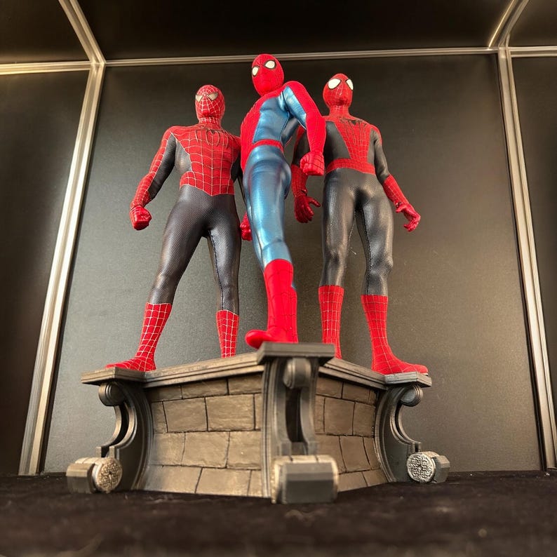 May include: Three red and blue Spider-Man action figures stand on a gray stone base. The figures are posed in different action stances.