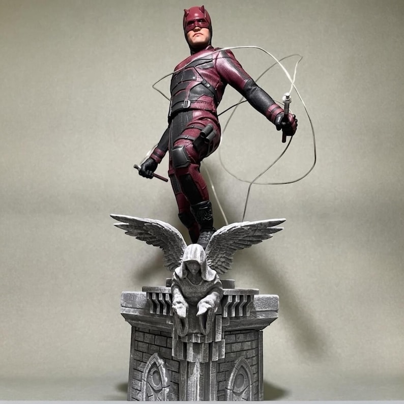 May include: A red and black Daredevil action figure stands on a gray stone base with a sculpted angel. The figure is holding a billy club and has a red mask and cape.