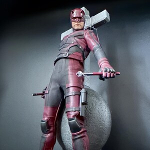 May include: A red and black action figure of Daredevil, a superhero from Marvel Comics, standing in front of a gray stone cross. The figure is holding a pair of batons in his hands.