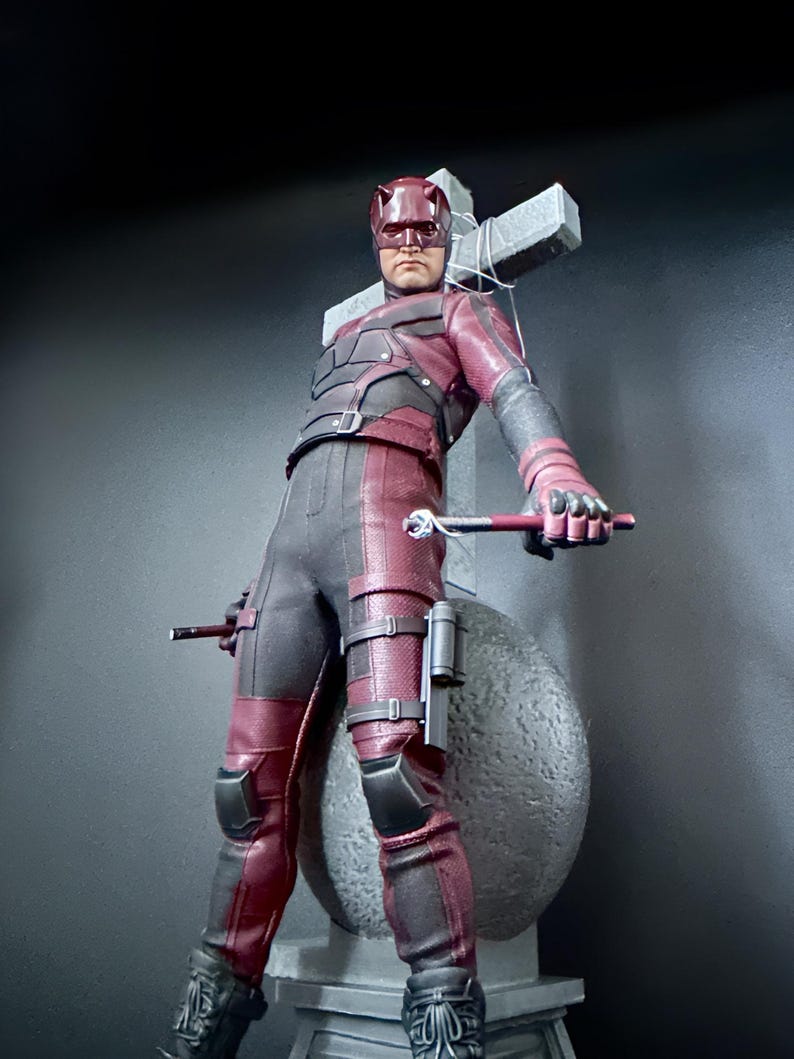 May include: A red and black action figure of Daredevil, a superhero from Marvel Comics, standing in front of a gray stone cross. The figure is holding a pair of batons in his hands.