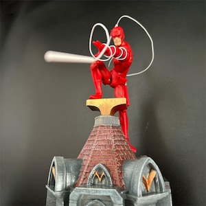 Può includere: Una action figure rossa di Daredevil in piedi su un tetto, con una corda bianca nelle mani. La figura è in una posa d'azione dinamica, con una gamba sollevata e le braccia distese. Il tetto è in pietra grigia e ha un camino in mattoni rossi.