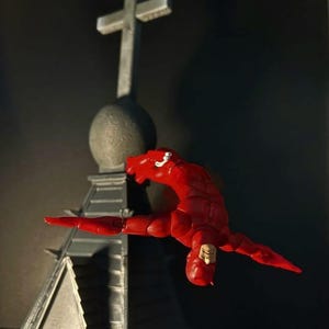 Può includere: Una action figure rossa in posa dinamica, apparentemente a mezz'aria, con braccia e gambe estese. La figura è posizionata di fronte a una struttura grigia sormontata da una croce. Lo sfondo è scuro.