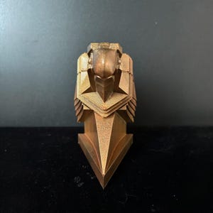 May include: A gold-colored, geometric, stylized sculpture of a bird's head with a sharp beak. The sculpture is made of a textured material and has a triangular base.