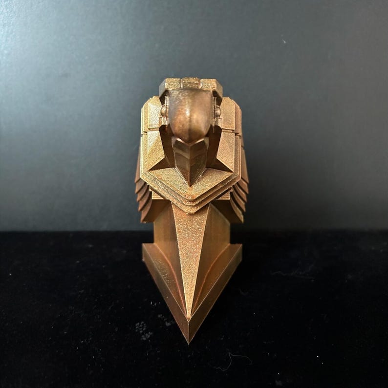 May include: A gold-colored, geometric, stylized sculpture of a bird's head with a sharp beak. The sculpture is made of a textured material and has a triangular base.
