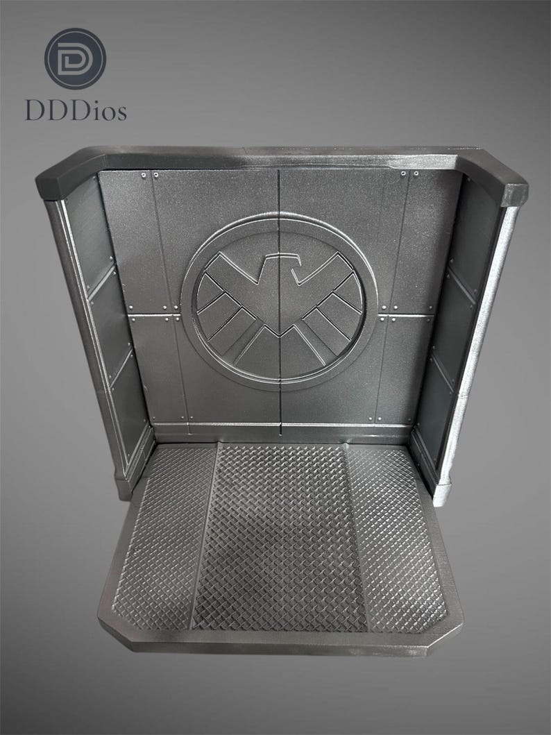May include: A silver-colored display stand with a textured floor and a circular emblem featuring a stylized bird. The stand has a rectangular shape with angled sides and the text "DDDios" in the upper left corner.