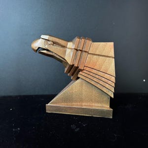 May include: A gold-colored eagle sculpture with a geometric design. The eagle's head is turned to the side, and its wings are spread out. The sculpture is mounted on a square base.